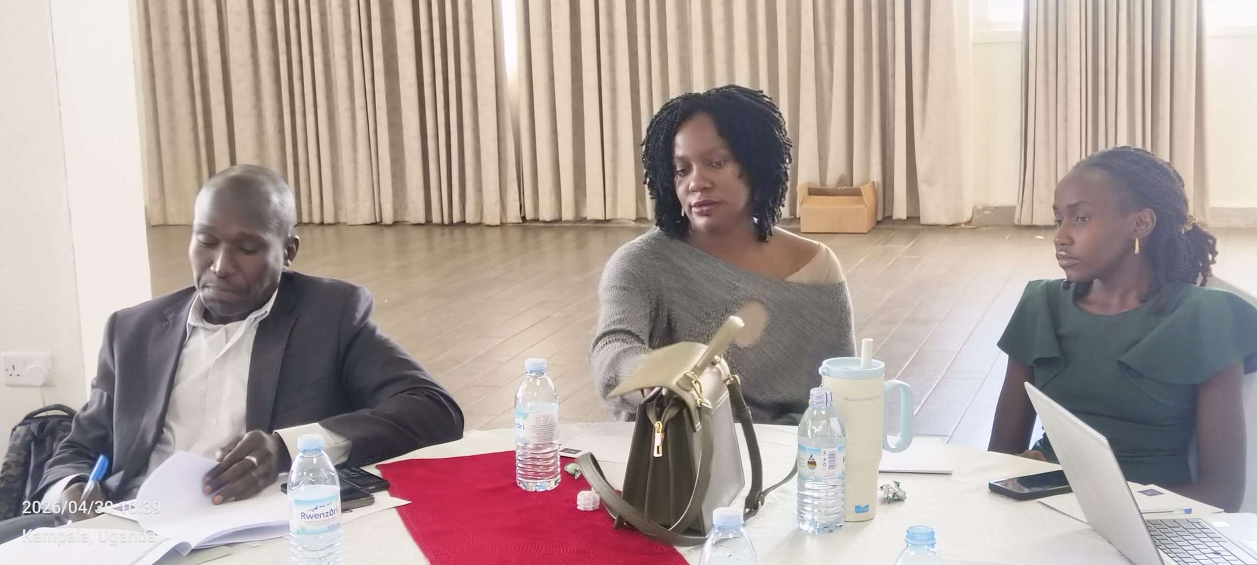 Three adults seated at a table in a conference room, reviewing papers while a woman in a gray sweater gestures toward documents; water bottles and a laptop on the table.