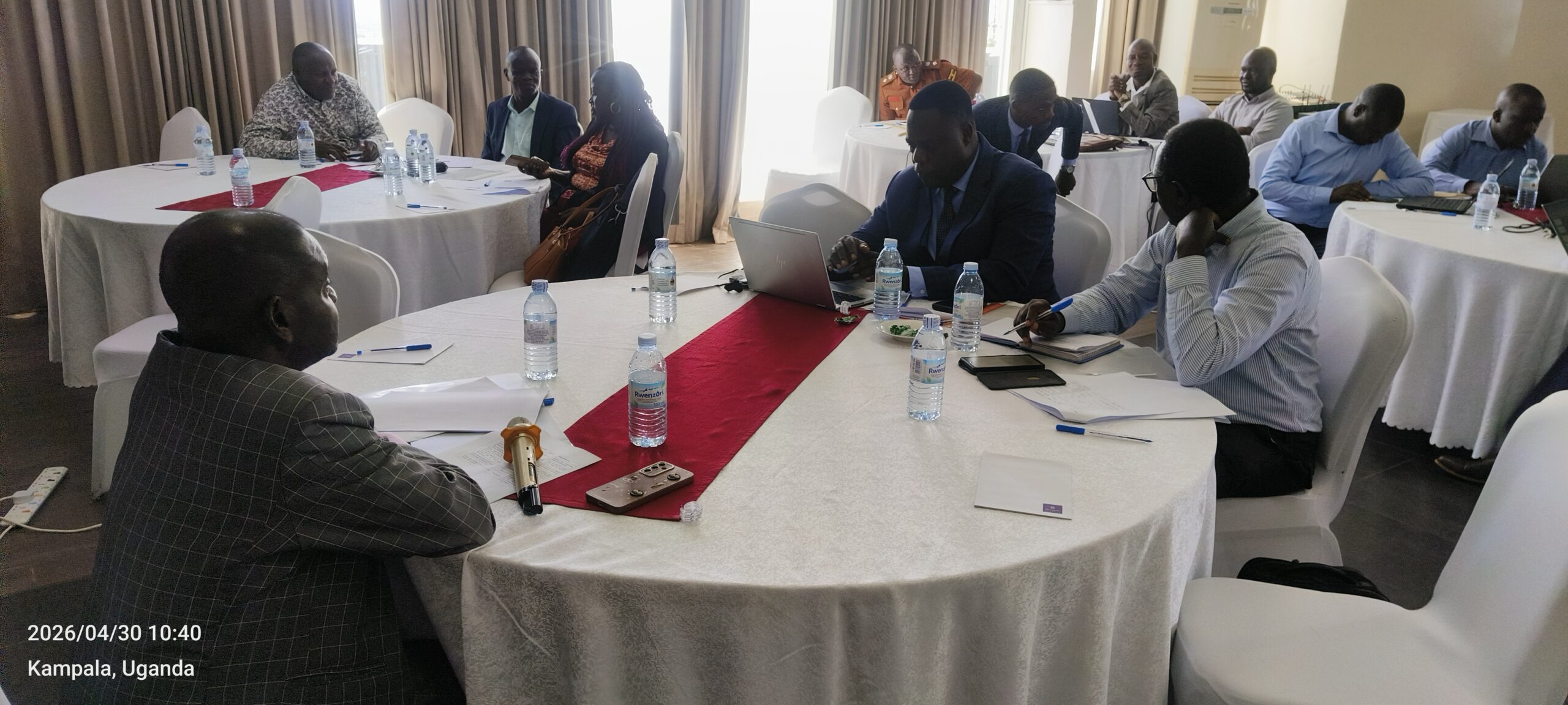 People in a conference room seated around round tables with laptops, papers, and water bottles during a meeting in Kampala, Uganda (date stamp visible).
