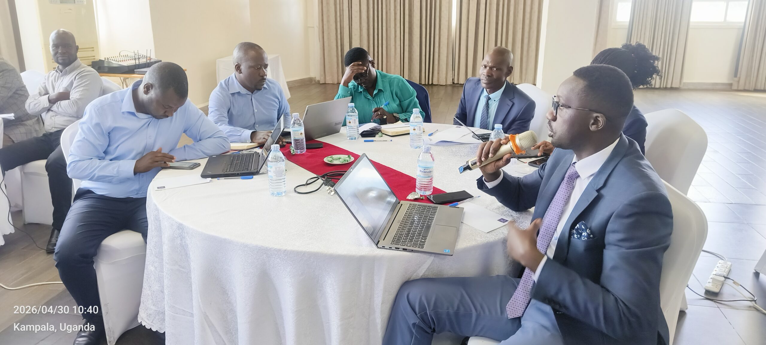 Six professionals around a round table with laptops; a man in a suit speaks into a microphone.