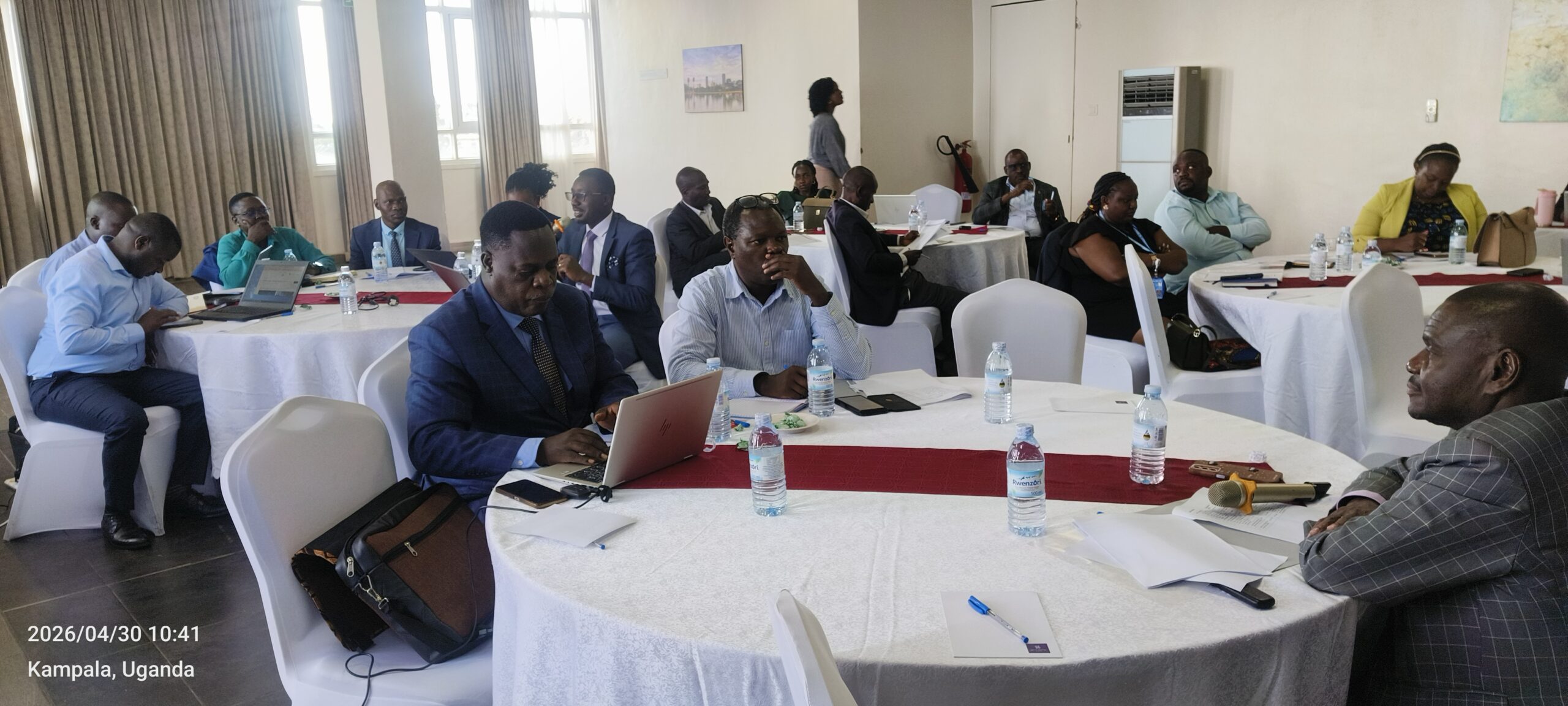 Professionals seated around round tables in a conference room, some with laptops, water bottles, and papers, in Kampala, Uganda (2026/04/30 10:41).