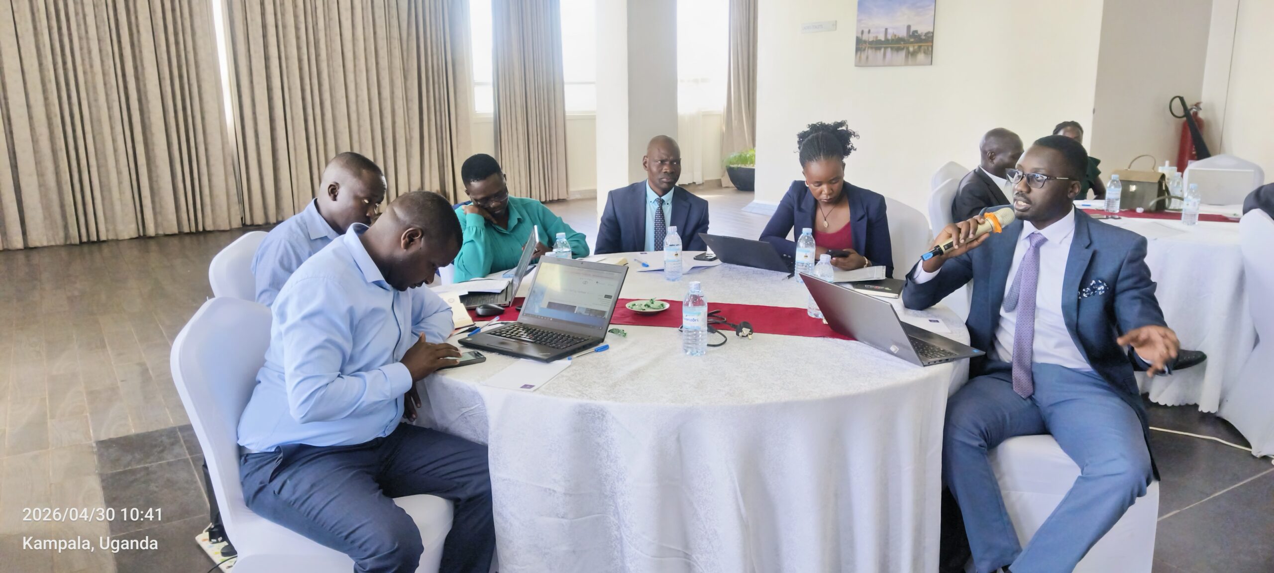 Six professionals in a round-table meeting with laptops; a man in a suit speaks into a handheld microphone.