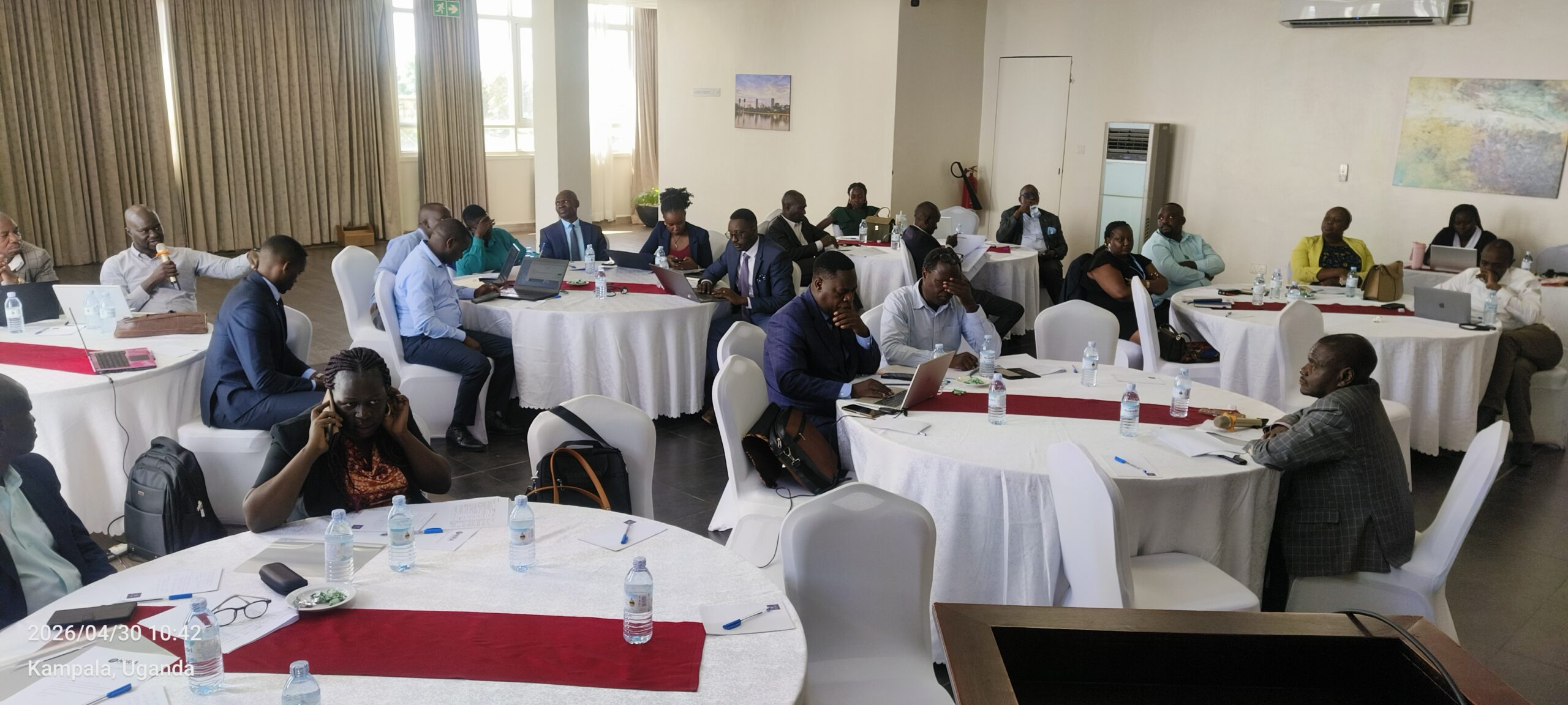 Professionals in formal attire seated around round tables in a bright conference room, laptops and papers in use during a meeting at a workshop in Kampala, Uganda.