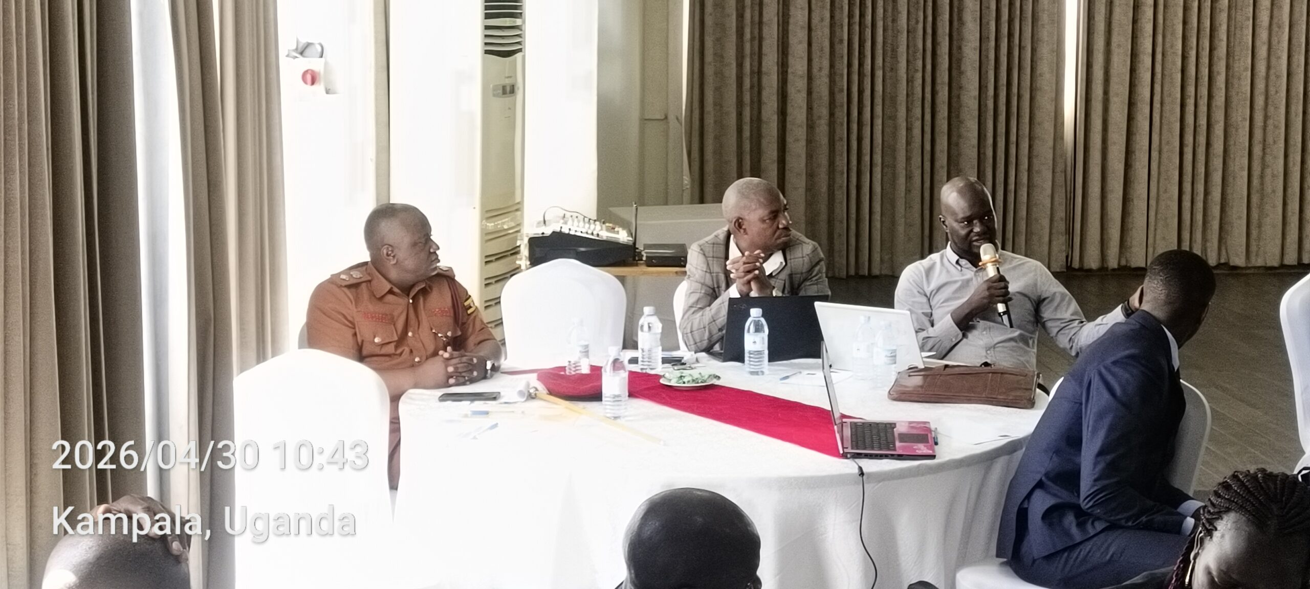 Panel discussion in a Kampala conference room: four men sit around a round table, one man speaks into a microphone while others listen. Laptops, water bottles, and a red table runner are visible.