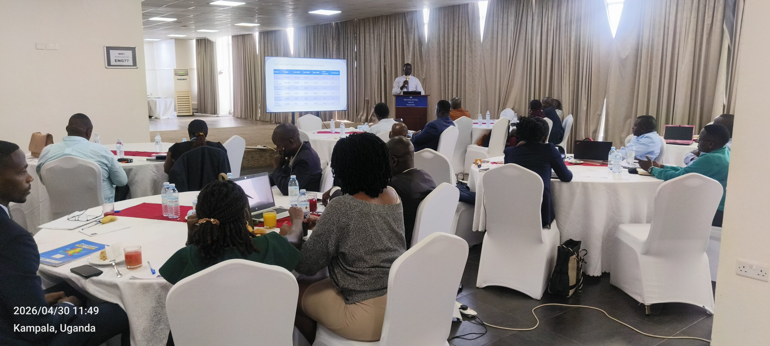 Conference room seminar: speaker at a podium presenting to round-table attendees with a projector screen behind him; Kampala, Uganda shown on the image.