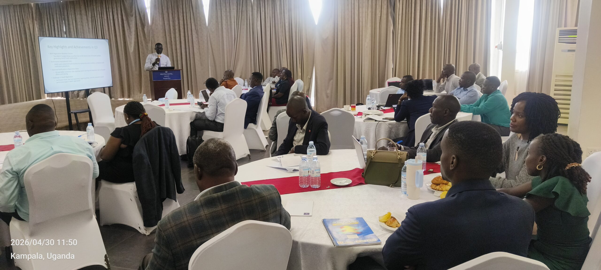 Business conference scene with a speaker at a podium addressing a room of attendees seated at round tables in a hotel banquet hall in Kampala, Uganda.