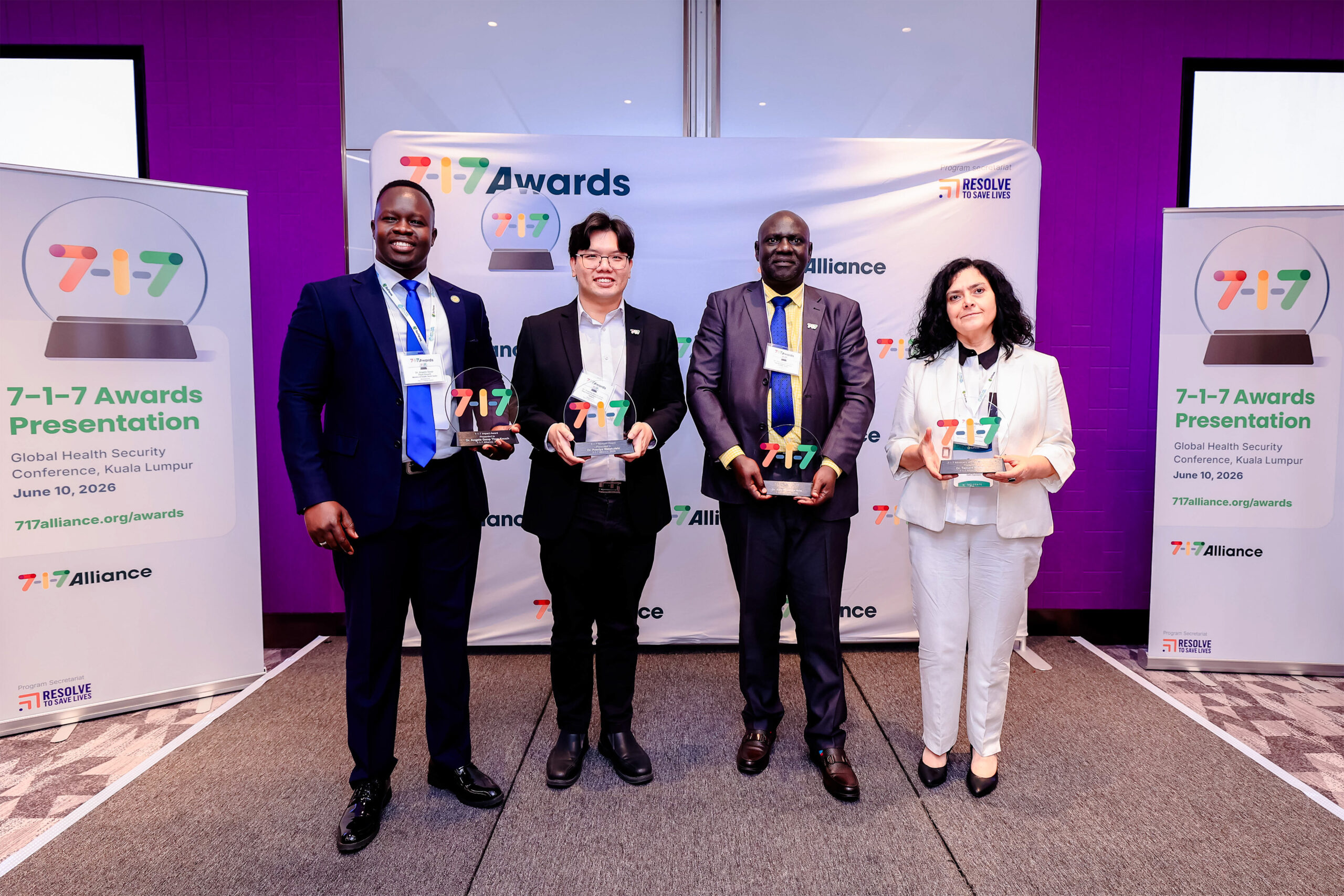Four professionally dressed individuals stand together on a stage holding glass trophies at an awards presentation, with 7-i7 Alliance banners in the background.