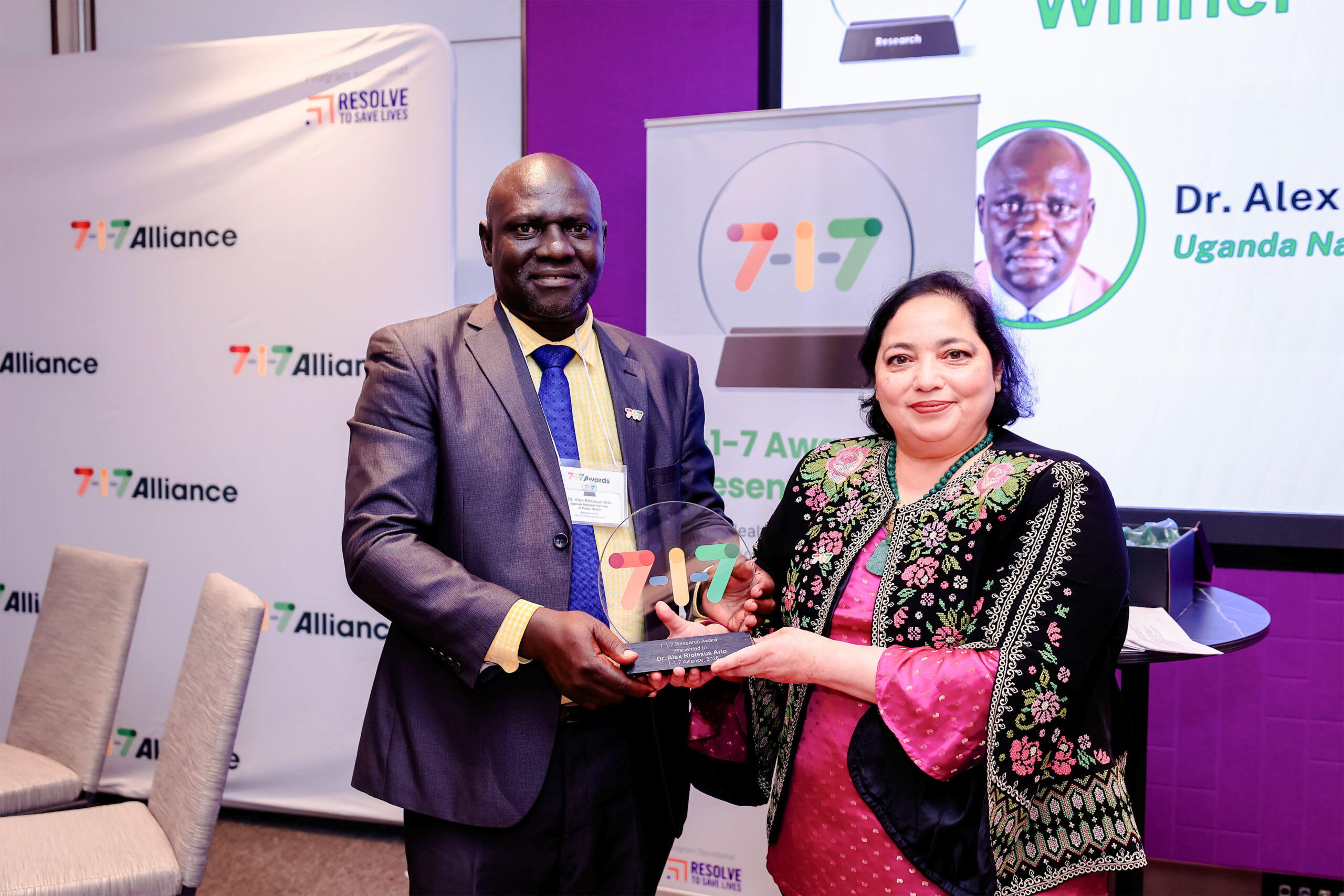 Man in a gray suit and blue tie presents a glass award to a woman in a pink patterned dress at a 7-7 Alliance event, smiling in front of banners behind them.